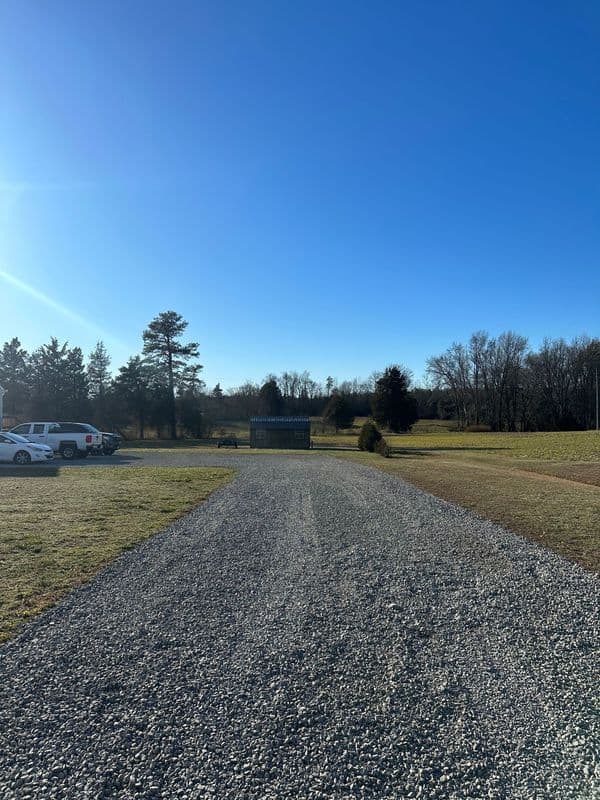 Gravel road leading to a sign, surrounded by trees under a clear blue sky.