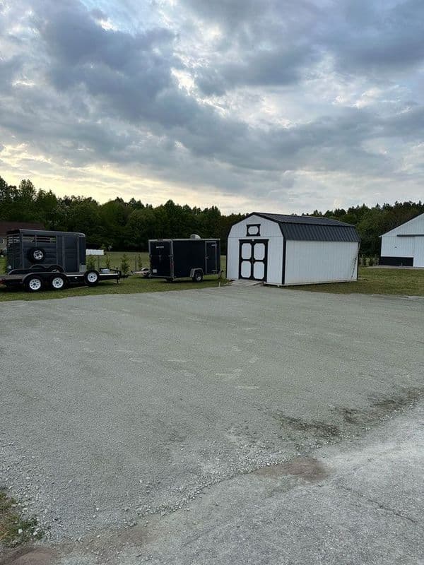 Gravel lot with trailers and a white shed under a cloudy sky in a rural setting.