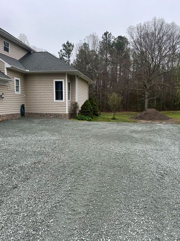 Gravel driveway beside a house with trees and a mound of soil in the background.
