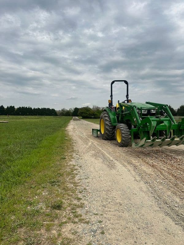 Green tractor plowing a gravel road in a rural landscape with overcast skies.