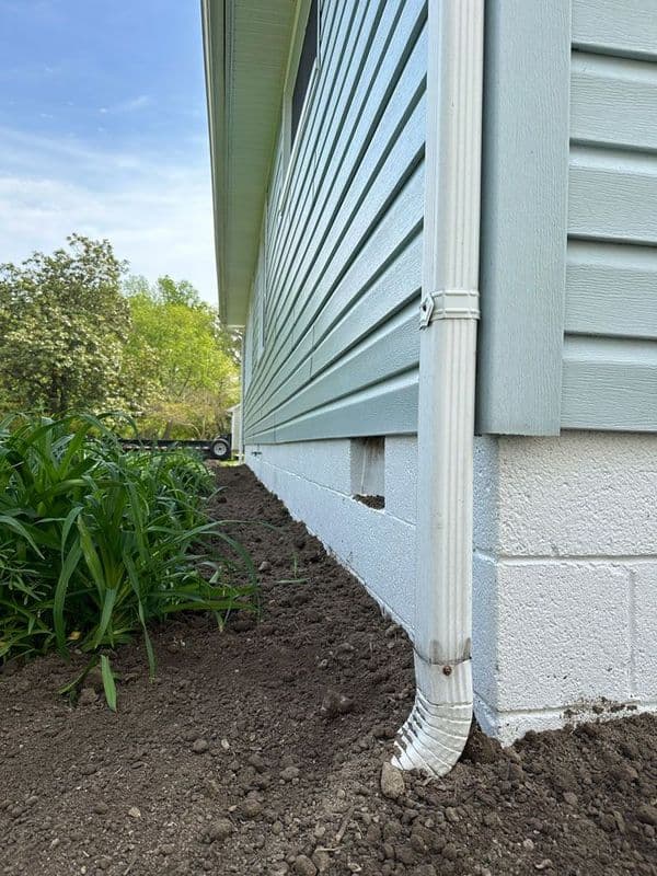 Downspout draining water from the house foundation, surrounded by fresh soil and greenery.
