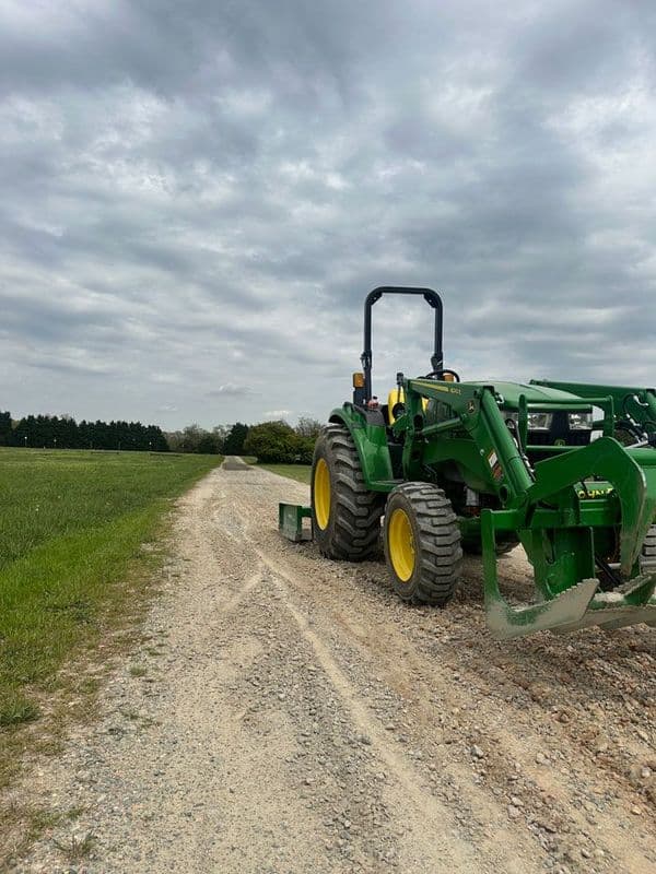 Green tractor on a gravel road surrounded by lush fields under a cloudy sky.