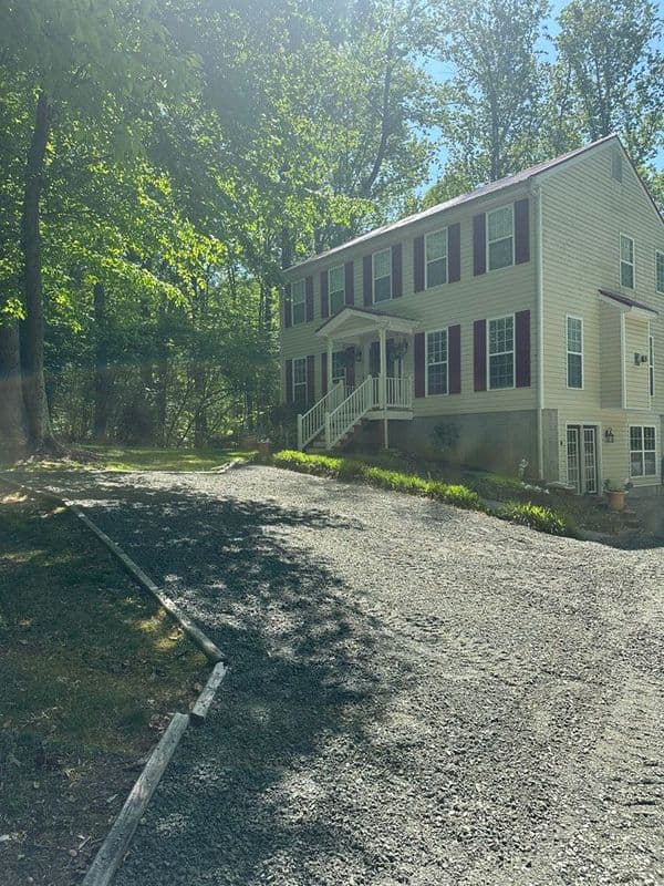 Two-story house with a gravel driveway surrounded by green trees and sunlight.