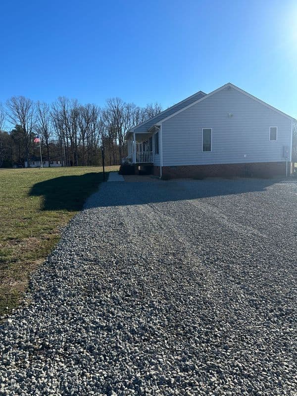 Gravel driveway leading to a light blue house surrounded by trees on a sunny day.