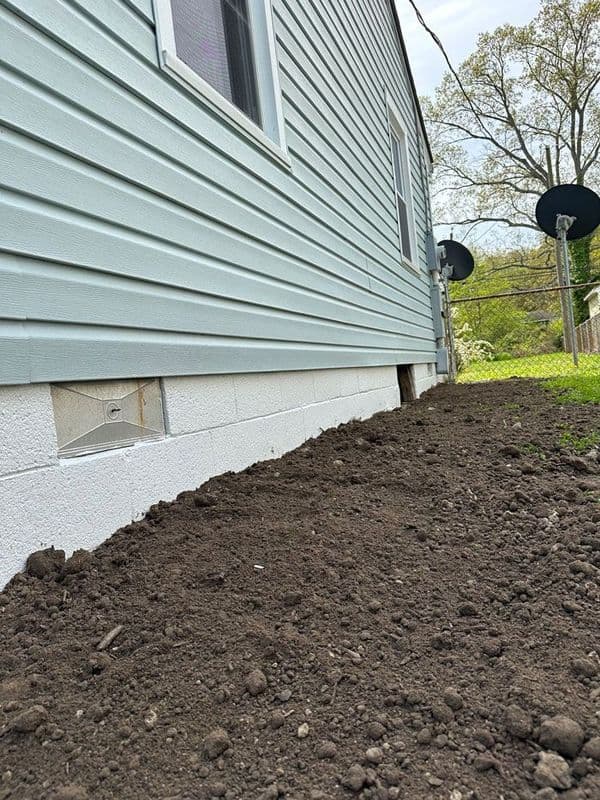 Freshly dug soil and foundation wall of a light blue-sided house with satellite dishes nearby.