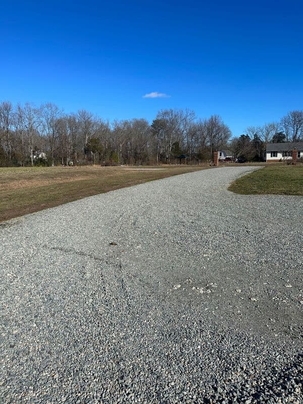 Gravel driveway leading to a house, surrounded by trees under a clear blue sky.