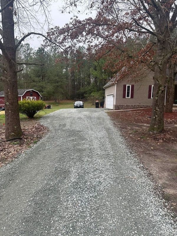Gravel driveway leading to a house surrounded by trees and outbuildings in a rural setting.