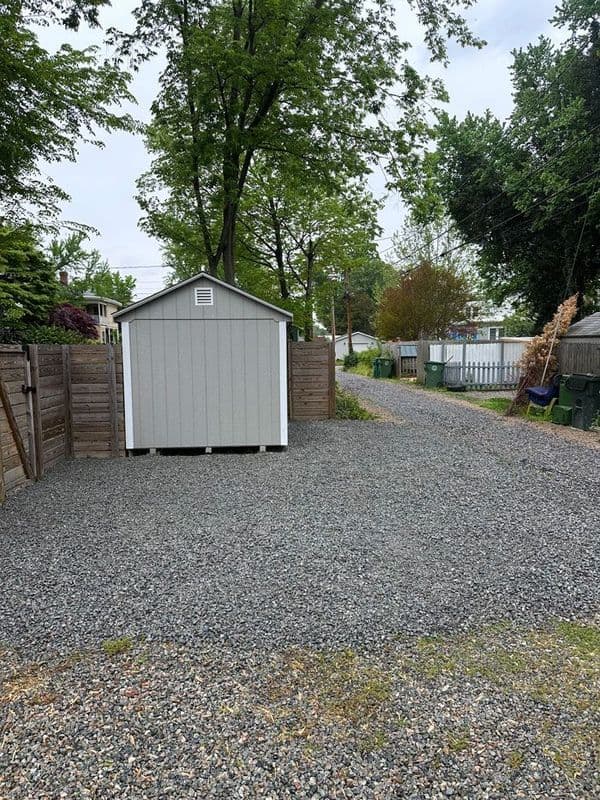 Gravel driveway with gray shed surrounded by trees and wooden fences.