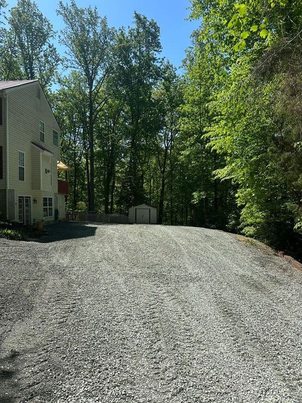 Gravel driveway beside a house, surrounded by lush green trees in bright sunlight.