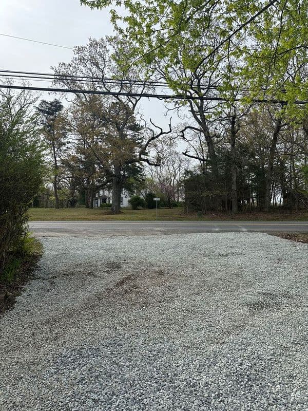 Gravel driveway leading to a residential area with trees and a road sign in the distance.