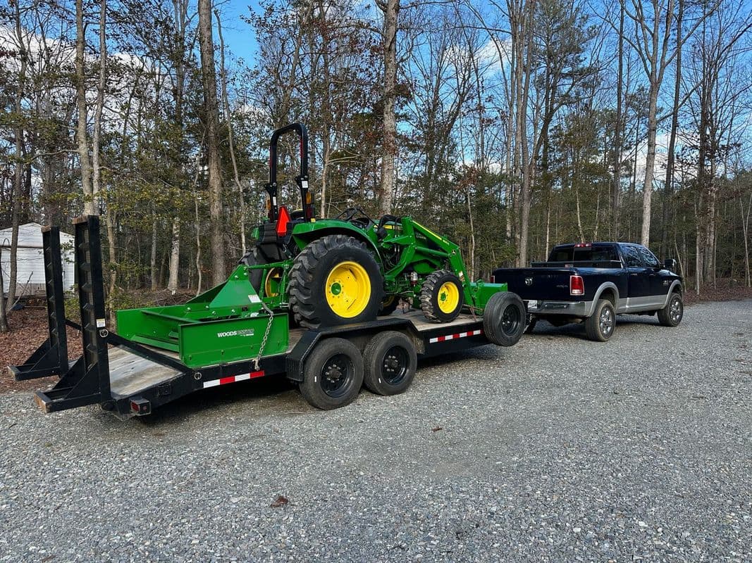 Green tractor and equipment on trailer hitched to black pickup truck in forested area.