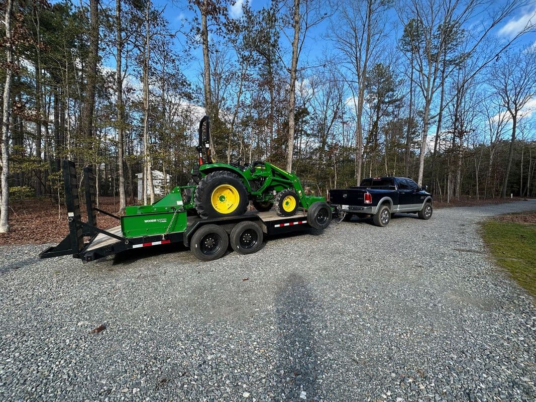 Green tractor on trailer towed by a black truck on a gravel road in a wooded area.