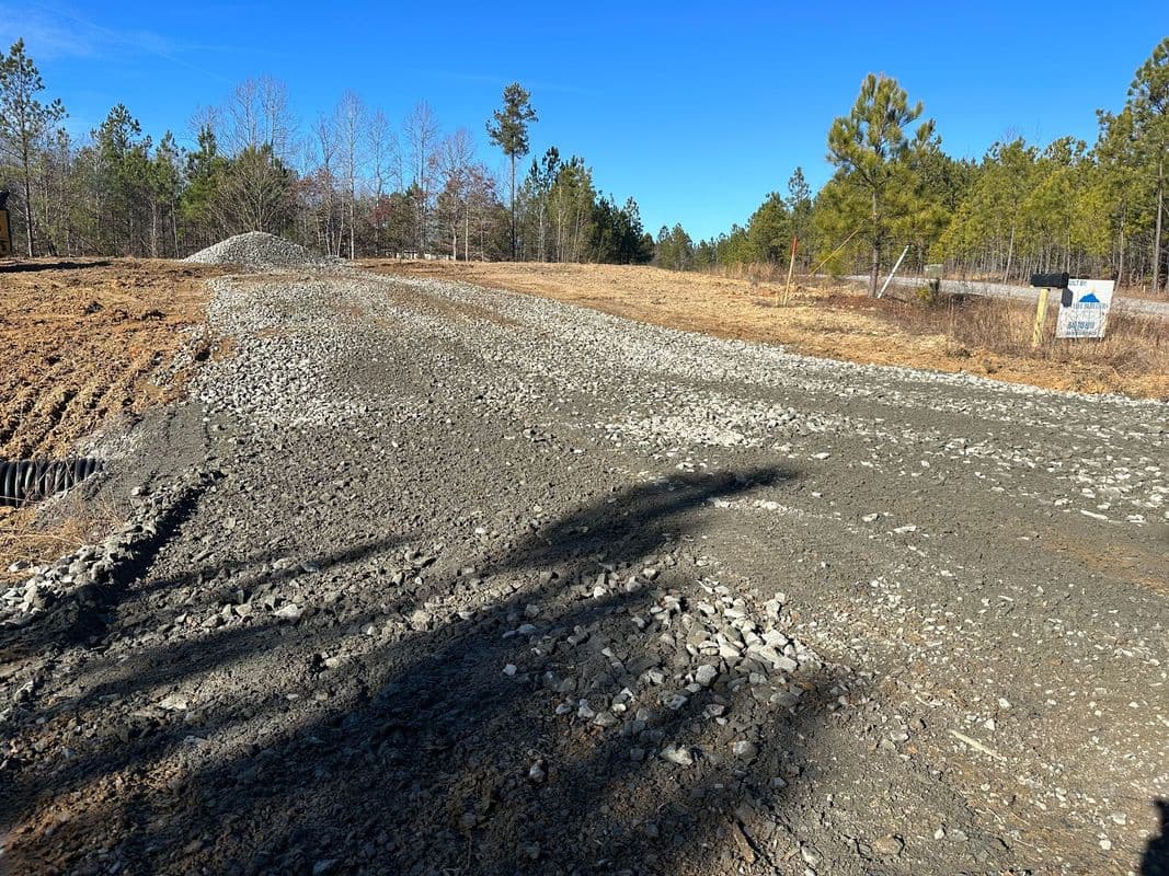 Gravel road under construction in a wooded area with clear blue sky and nearby sign.